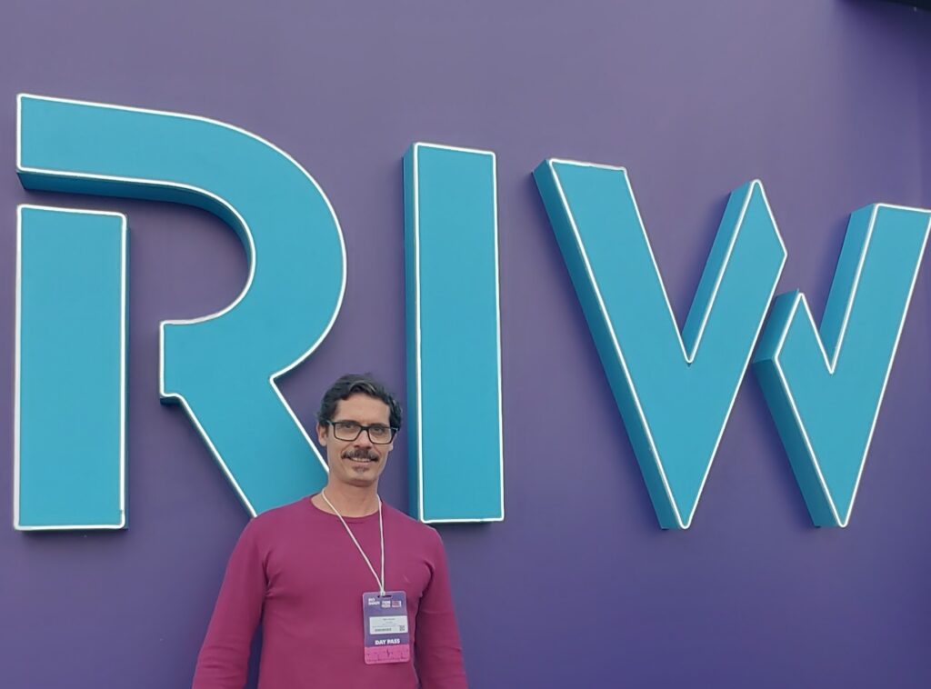 Um homem de óculos e bigode, com camisa bordô e crachá DAY PASS, sorri diante de um grande letreiro azul RIW em uma parede roxa.